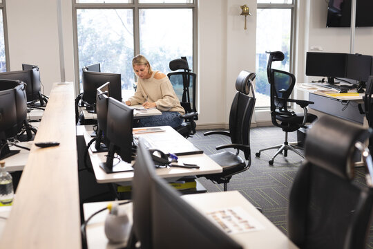 Woman writing in notebook at open-plan office desk with monitors keyboard mouse and ergonomic chair