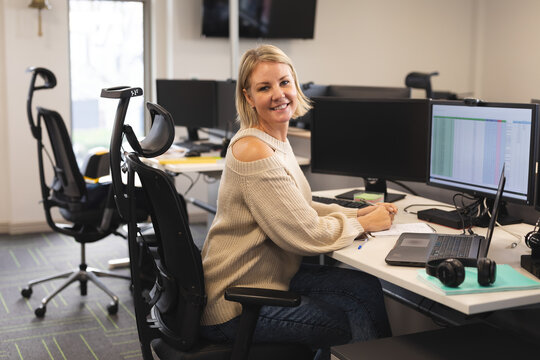 Woman typing on laptop at open-plan office desk, using dual monitors and over-ear headphones
