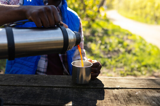 Fototapeta African American man pouring coffee from steel thermos into mug at wooden picnic table in park