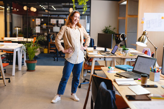Female office worker standing hands on hips in open-plan office, with laptops and potted plants