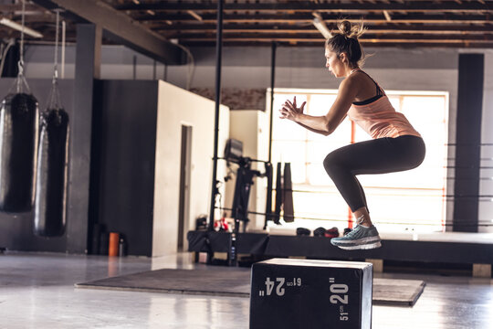 Young adult woman performing box jump at industrial gym with punching bags and barbells, copy space