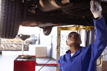 African American female mechanic inspecting undercarriage on lift in repair shop, copy space