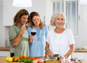 Cheerful couple and old woman drinking wine and talking with eath other while cooking in the kitchen at home