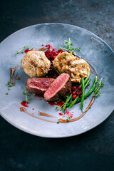 Traditionally roasted fillet of saddle of venison with bread dumplings, mushrooms and bean sticks served with lingonberry sauce as close-up on a design plate with a rustic background