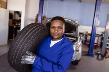 African American female mechanic lifting car tire over shoulder in repair garage, with tool chests