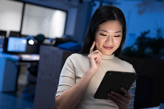 Asian woman tapping tablet in dimly lit modern open?plan office with computer monitors, copy space