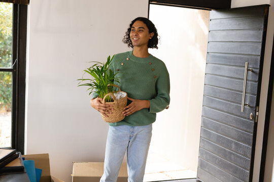 Non-binary person holding green potted plant in woven basket in entrance hallway, with moving boxes