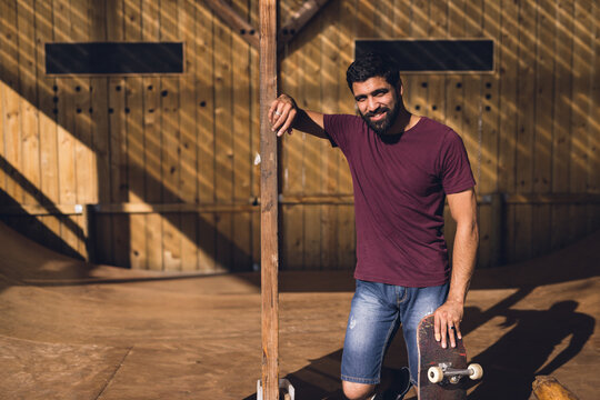 Man leaning on beam in indoor skate ramp, holding scratched skateboard, copy space