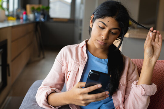 Middle Eastern teenage girl sitting on couch at home, twirling hair and looking at smartphone