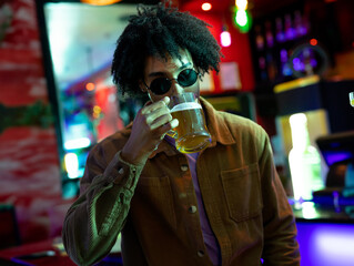 African American man drinking from glass mug of beer in bar interior, with neon lighting