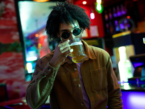 African American man drinking from glass mug of beer in bar interior, with neon lighting