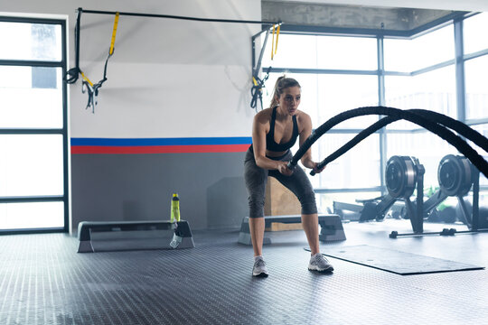 Woman performing battle ropes exercise in functional gym, with TRX straps, water bottle, copy space