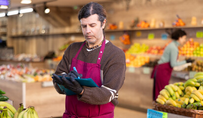 Adult man seller in apron conducting inventory with checklist in vegetable shop
