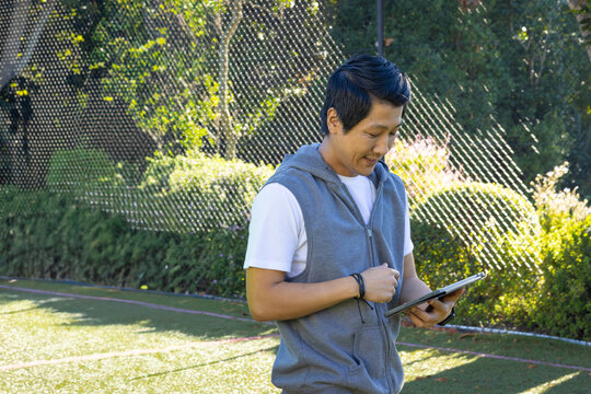 Mid-adult Asian man tapping tablet on green turf field by black net fence, copy space