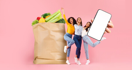Three friends joyfully pose in front of a large paper grocery bag filled with fresh produce,...