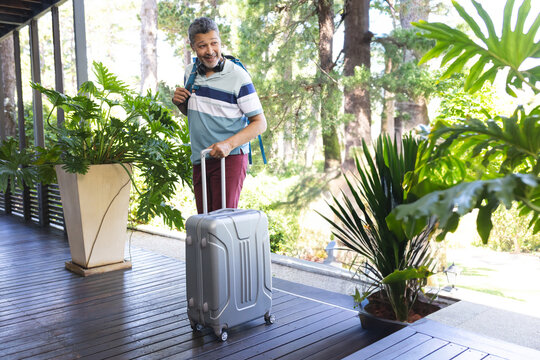 Man pulling silver rolling suitcase on shaded wooden veranda, carrying blue backpack and headphones