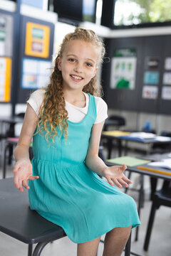 Female child sitting on student desk in school classroom, showing colorful notebooks and posters