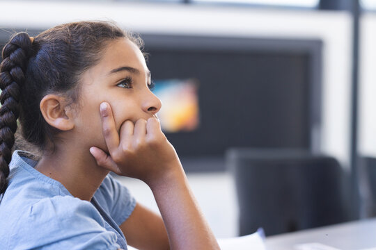 Girl child resting chin on hand, gazing right with notebook and screen in classroom, copy space