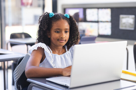Girl working on laptop at classroom desk with black chair, notebooks, pencils and striped backpack