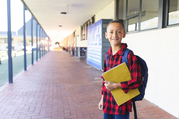 10-year-old boy standing in school hall, carrying yellow notebooks and blue backpack, copy space