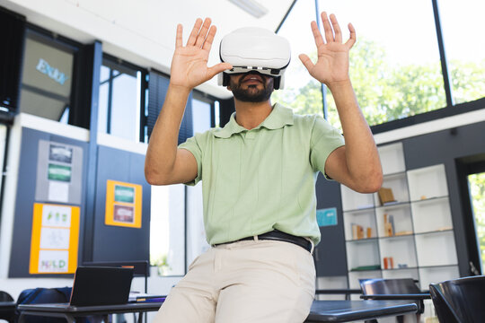 Young Asian Indian man sitting on table using VR headset in modern classroom, with laptop
