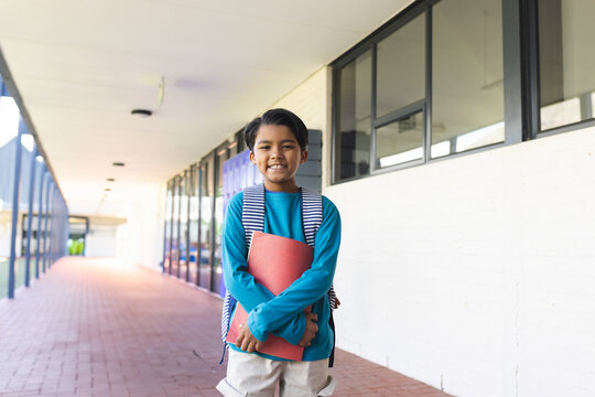 Boy student standing in covered school corridor smiling with red binder and striped backpack