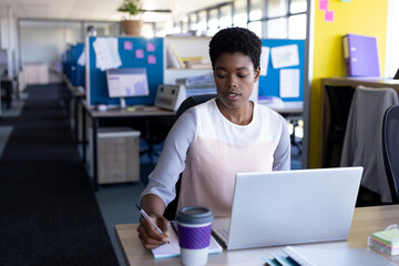 Young African American woman working on laptop at office desk, jotting notes in notepad, copy space