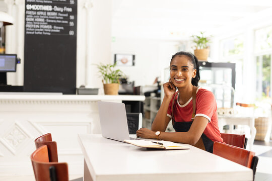 Asian Indian woman writing in open notebook at communal café table with laptop, copy space