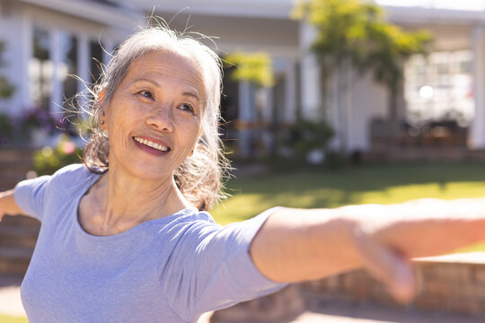Senior Asian woman stretching arms horizontally in front of sunny suburban house, with gentle smile