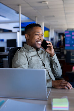 African American man sitting at desk in modern open-plan office, talking on smartphone with laptop