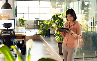 Young Asian Korean woman using tablet by glass partition with notes in open-plan office, copy space