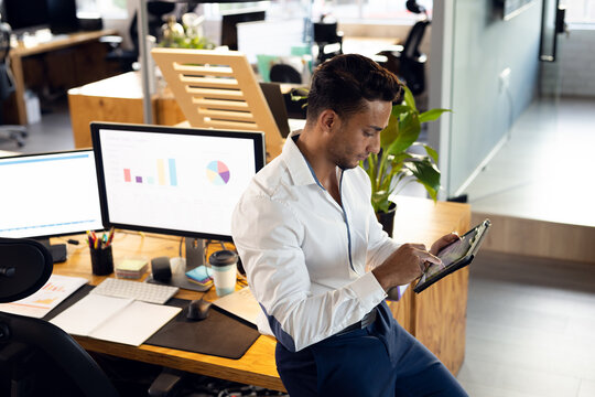 Young adult Hispanic male leaning on desk in modern office, viewing tablet and charts, copy space