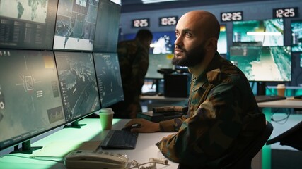 Military soldier in mission control center overseeing national safety operations with devices,...