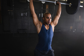 Man performing overhead barbell lift in dimly lit gym, with weight racks and rubber flooring
