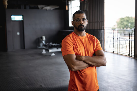 Mid-adult Multiracial man standing with arms crossed in gym with dumbbell rack, exuding focus