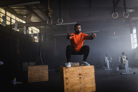 Man performing box squat on wooden plyo box in warehouse gym, including rings and rowing machines