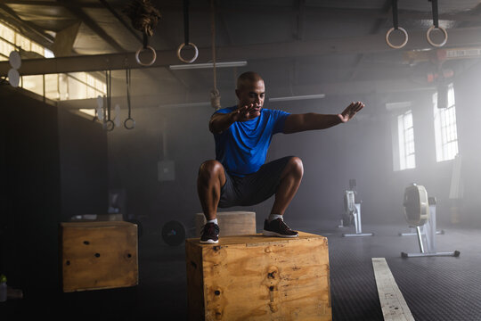 Multiracial man squatting on plyometric box in industrial gym, showcasing strength training