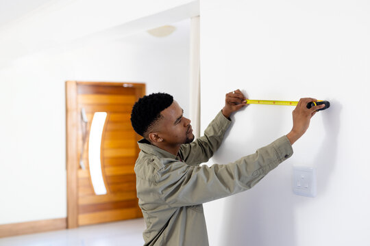 African American man measuring white wall in bright residential entryway, with yellow tape measure