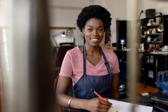 African American woman sketching on workbench in makerspace, holding pencil over open sketchbook