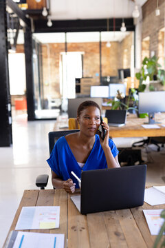 Young African American woman talking on smartphone and using laptop at open?plan office desk