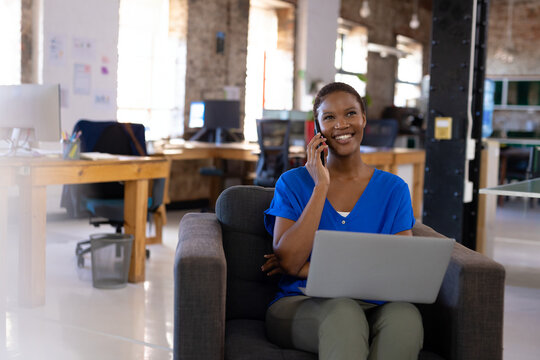 African American woman talking on smartphone while using laptop in armchair in office, doing work