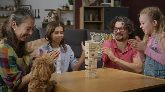 Medium shot of laughing Caucasian mother, father with limb difference, young son and daughter playing wooden blocks game at home, cheering and clapping, pet dog jumping around table