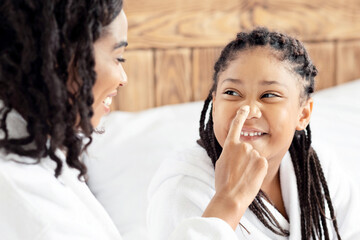Young African Mom And Her Little Daughter In Bathrobes Having Fun And Laughing At Home, Relaxing On Bed After Beauty Routine In Bathroom, Loving Black Mother Tenderly Touching Child's Nose, Closeup
