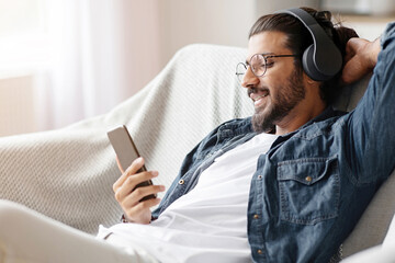 Relaxed indian guy listening music on smartphone with wireless headphones, relaxing on couch at home, enjoying favorite songs on mobile phone, downoading playlist, resting on comfy sofa in living room