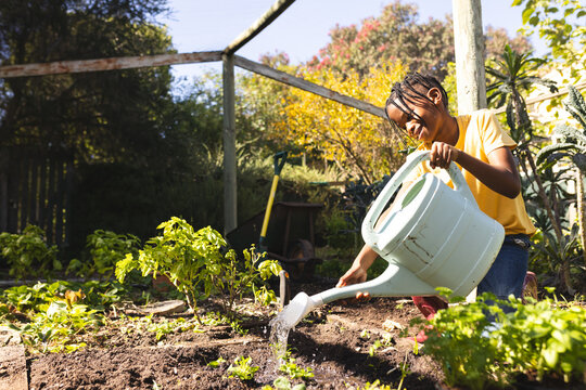 Young African American girl kneeling in home garden, watering seedlings with green can, copy space