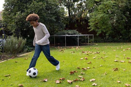 Boy kicking black and white soccer ball in backyard, with trampoline and fallen leaves, copy space