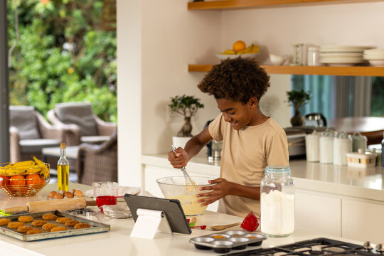 Boy whisking batter in glass bowl at modern home kitchen island, using tablet recipe, copy space - Powered by Adobe