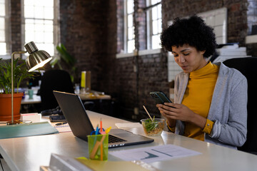 Woman checking smartphone at desk in loft office with laptop and salad bowl, copy space