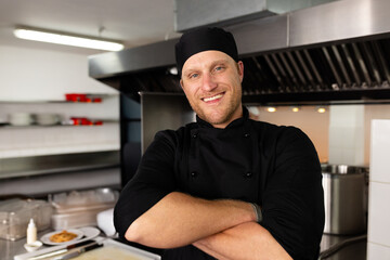 Male chef standing with folded arms in commercial kitchen showing cooking hood and shelves