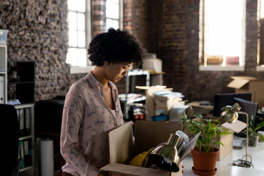 Young woman packing cardboard box in industrial office loft, with metal desk lamp and potted plant